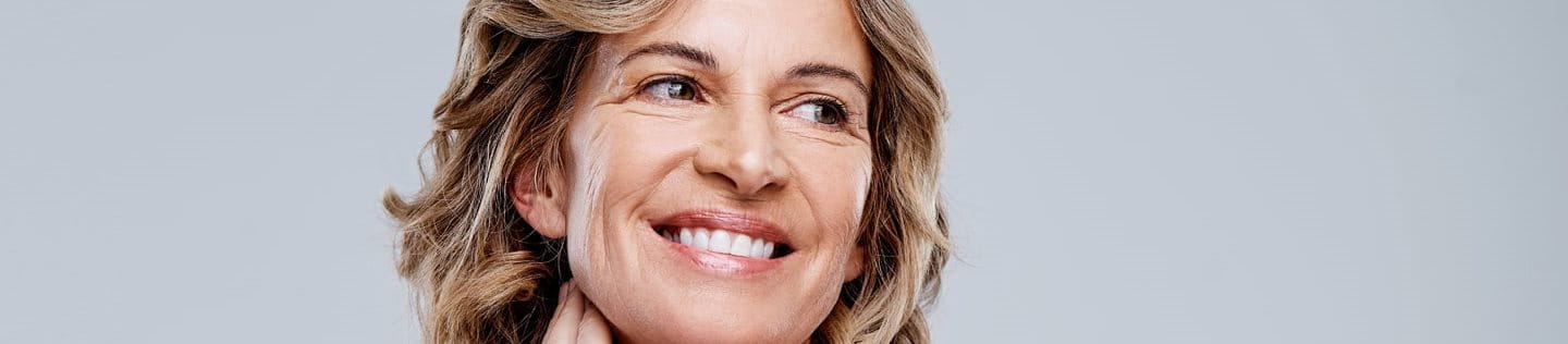 Uśmiechnięta, dojrzała kobieta o krótkich, falujących blond włosach delikatnie dotyka boku swojej szyi, patrząc poza kadr. Ma odkryte ramiona, a jej naturalna, promienna cera wyróżnia się na jasnoszarym tle.