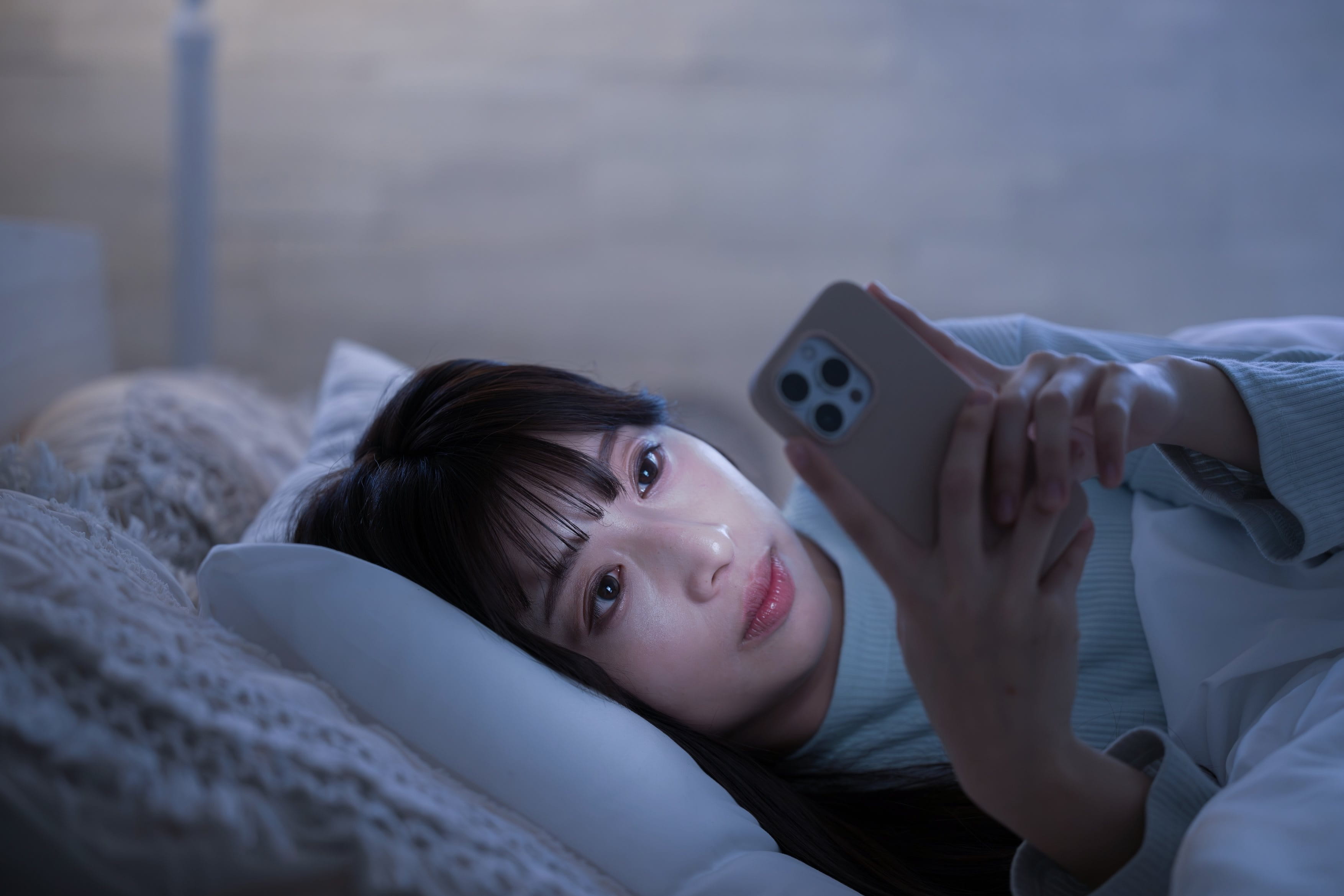 Eine junge Frau liegt in einem abgedunkelten Zimmer auf einem Bett und schaut auf das helle Display ihres Smartphones