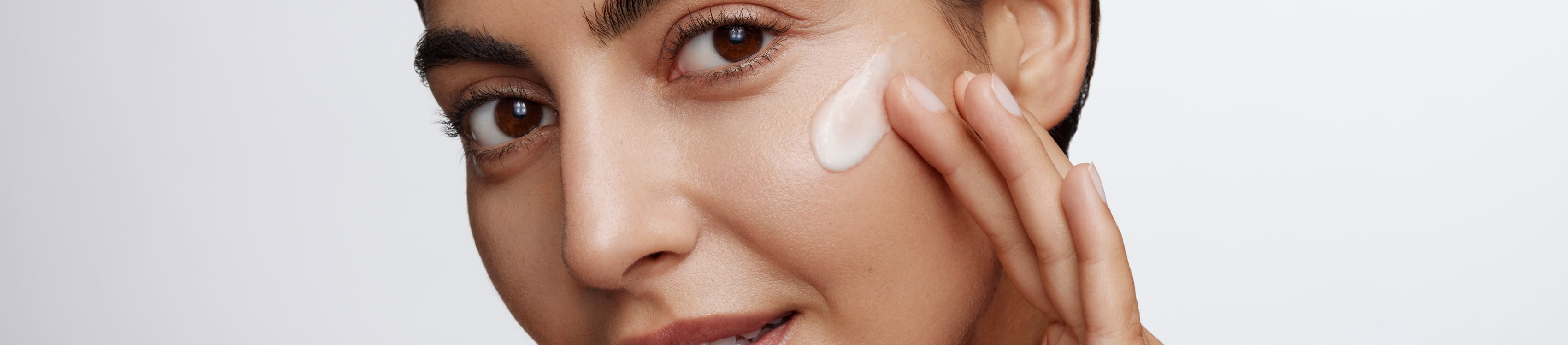 Mulher aplicando hidratante facial na bochecha, com um visual de maquiagem natural e pele suave e radiante.