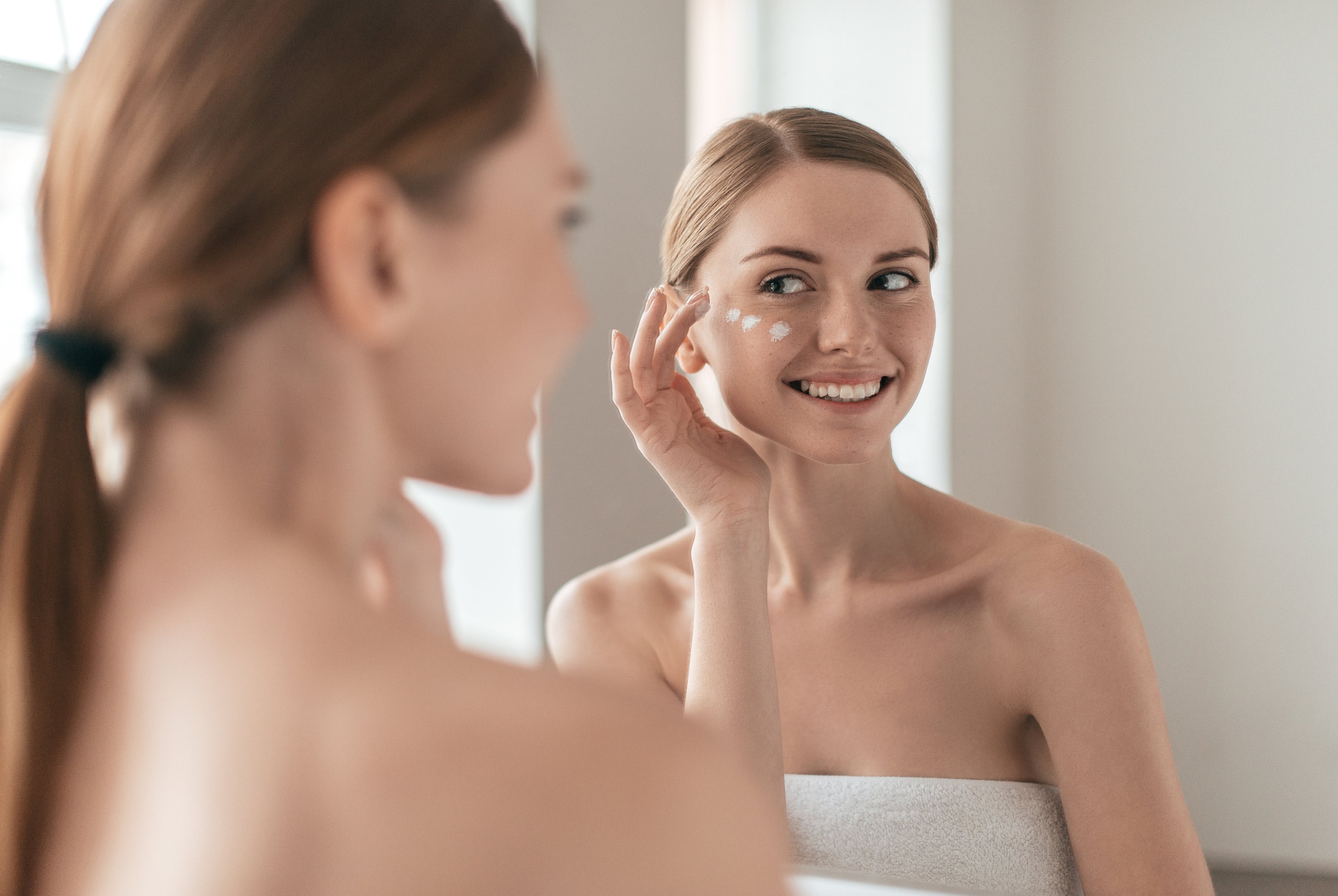 Eine junge Frau mit Zopf und in ein Handtuch gewickelt schaut in den Spiegel. Sie trägt mit einer Hand Creme auf ihre Wangenpartie auf. Der andere Arm ist nach unten gestreckt. Auf der linken Seite sieht man verschwommen ihren Umriss von hinten, während das Spiegelbild fokussiert ist.