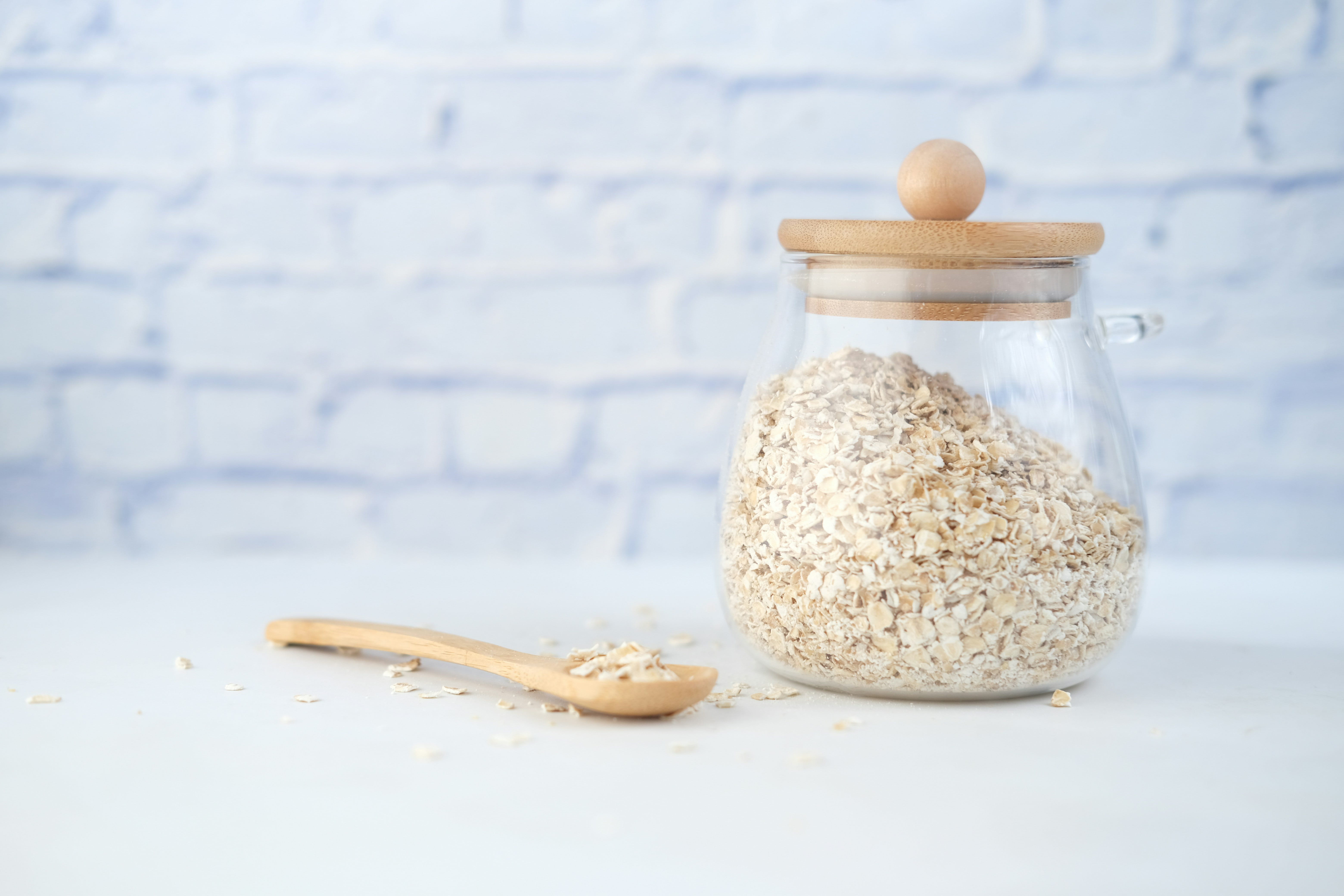 Ein mit Haferflocken gefüllter Glasbehälter mit Holzdeckel steht auf einem weißen Untergrund vor einer weiß verputzten Wand. Daneben liegt ein Holzlöffel mit Haferflocken, einige davon lose um den Löffel verteilt.