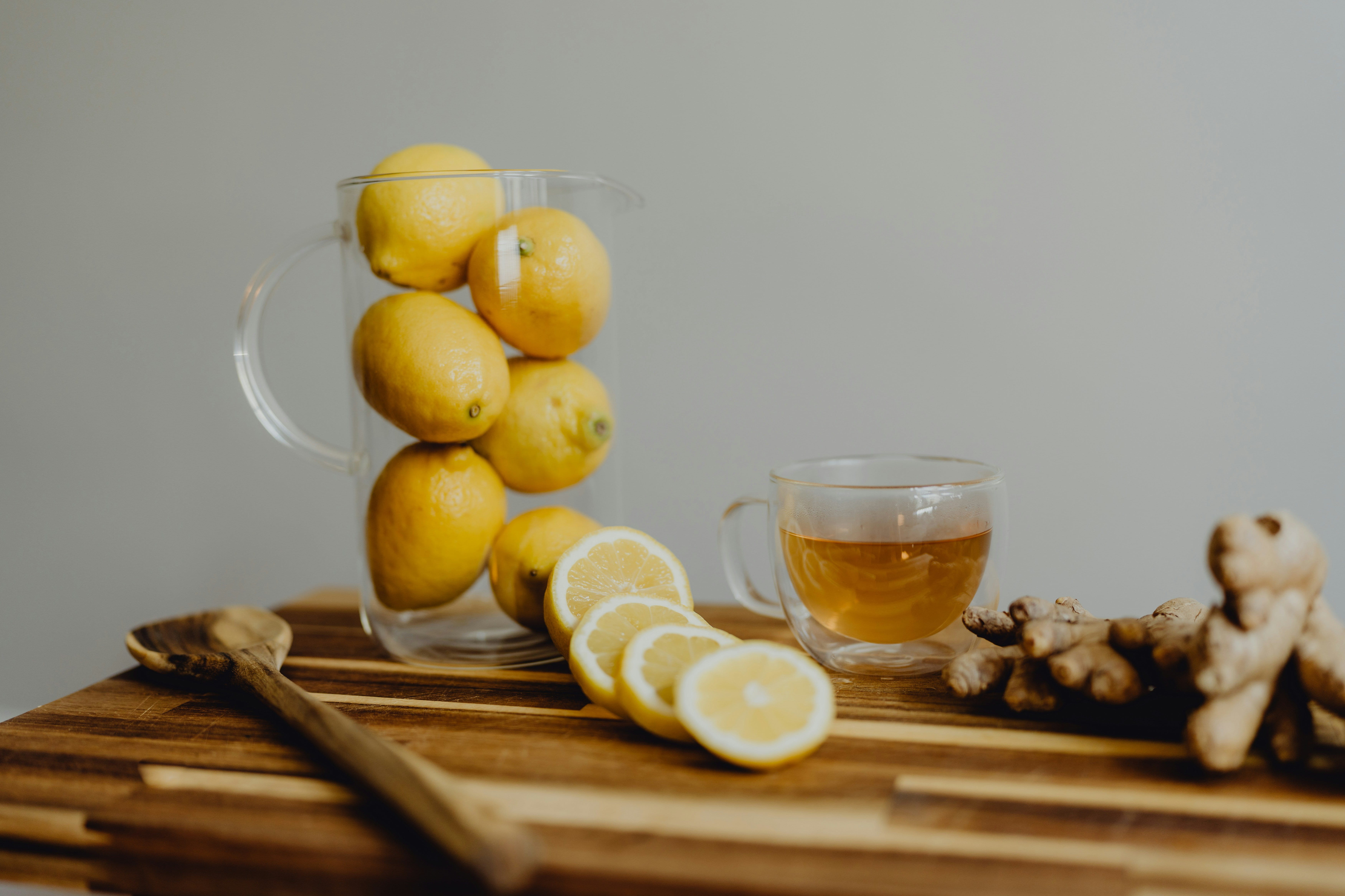 Eine Holztischplatte mit einem Glass Zitronen, von denen einige aufgeschnitten auf dem Tisch liegen, einer Tasse mit einer hellen Flüssigkeit und Ingwer