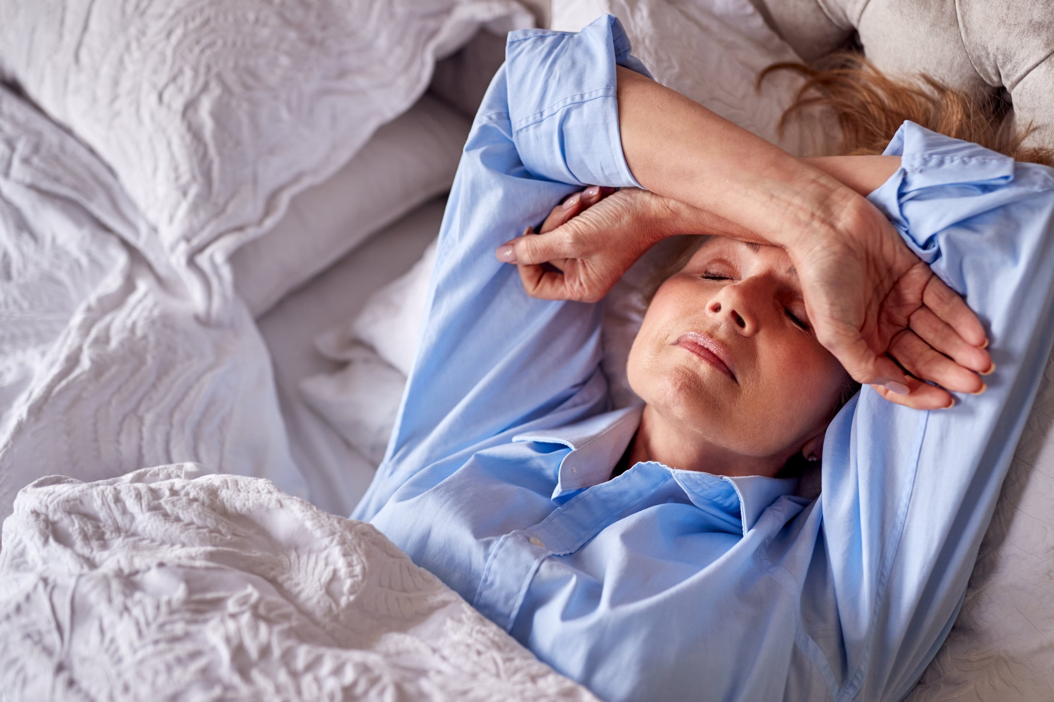 Eine ältere Frau in einem blauen Hemd liegt im Bett, hat die Augen geschlossen und legt ihre Arme über die Stirn, während sie entspannt ruht.