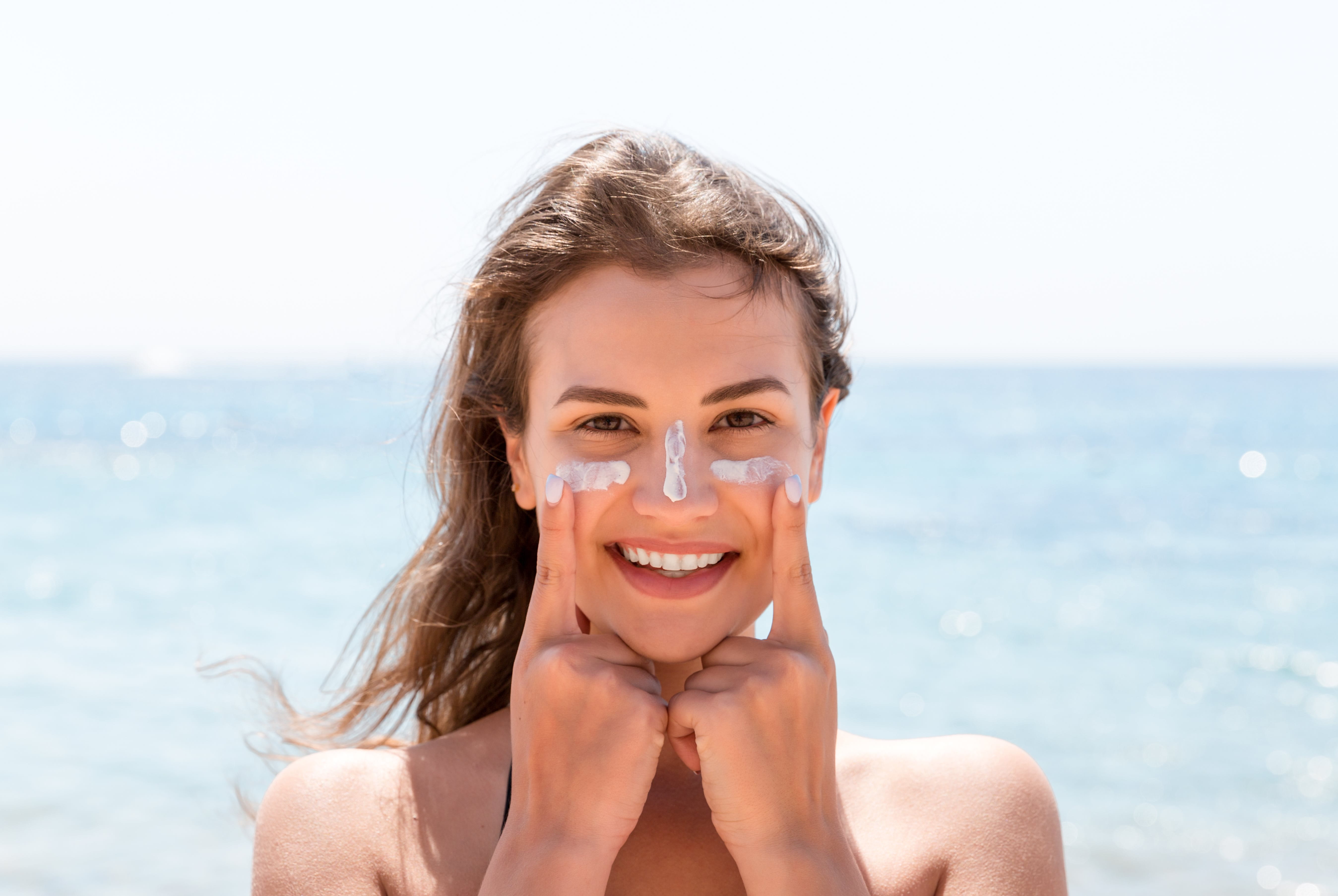 Frau am Strand trägt Sonnencreme auf Gesicht auf und lächelt in die Kamera.