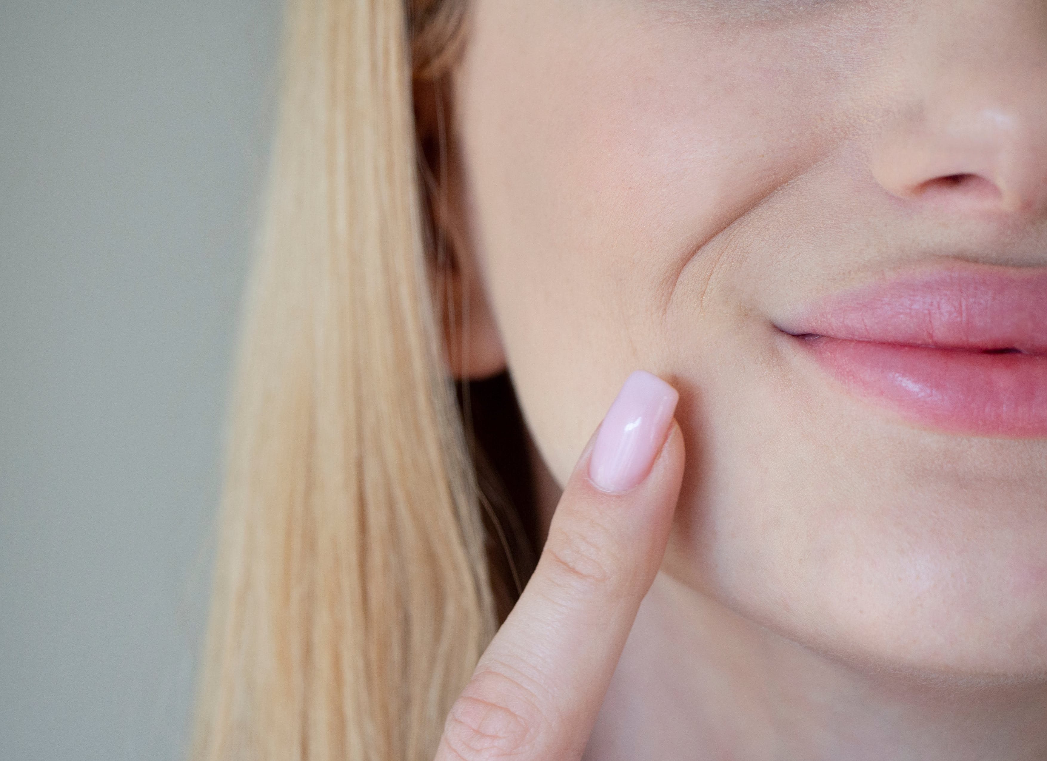 Eine Frau mit blonden Haaren und gepflegten Nägeln zeigt mit ihrem Finger auf eine feine Nasolabialfalte neben ihrem Mund, während sie leicht lächelt.