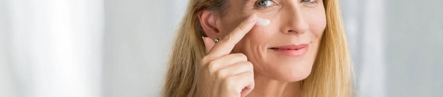 Eine lächelnde Frau mit langen blonden Haaren trägt eine Feuchtigkeitscreme auf ihre Wange auf. Sie blickt freundlich in die Kamera, während sie ihre Hautpflege-Routine durchführt.