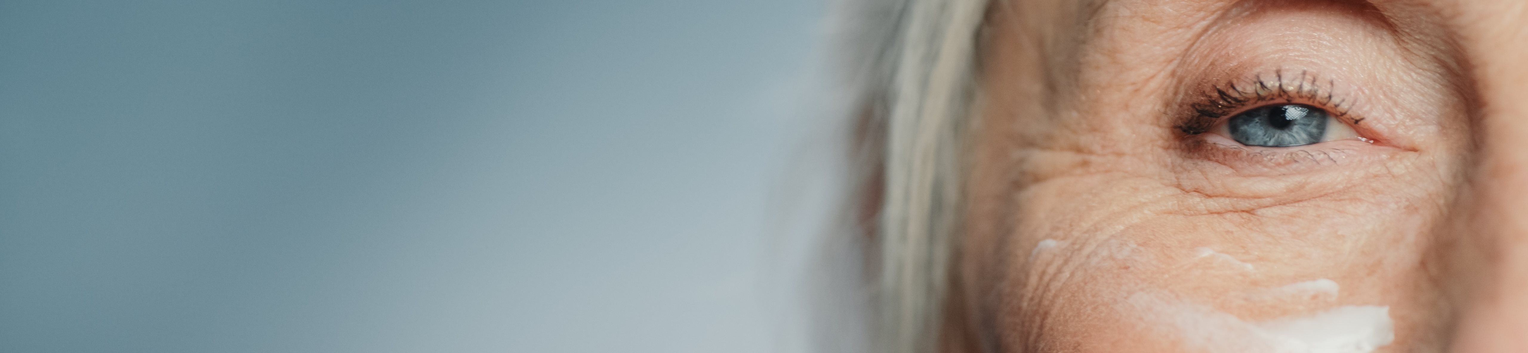 Nahaufnahme der Gesichtshälfte einer lächelnden älteren Frau mit sichtbaren Falten, die sich Creme auf die Wange aufträgt