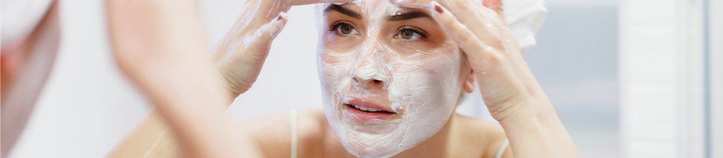 Woman applying a facial mask to her face while looking in a mirror during her skincare routine.