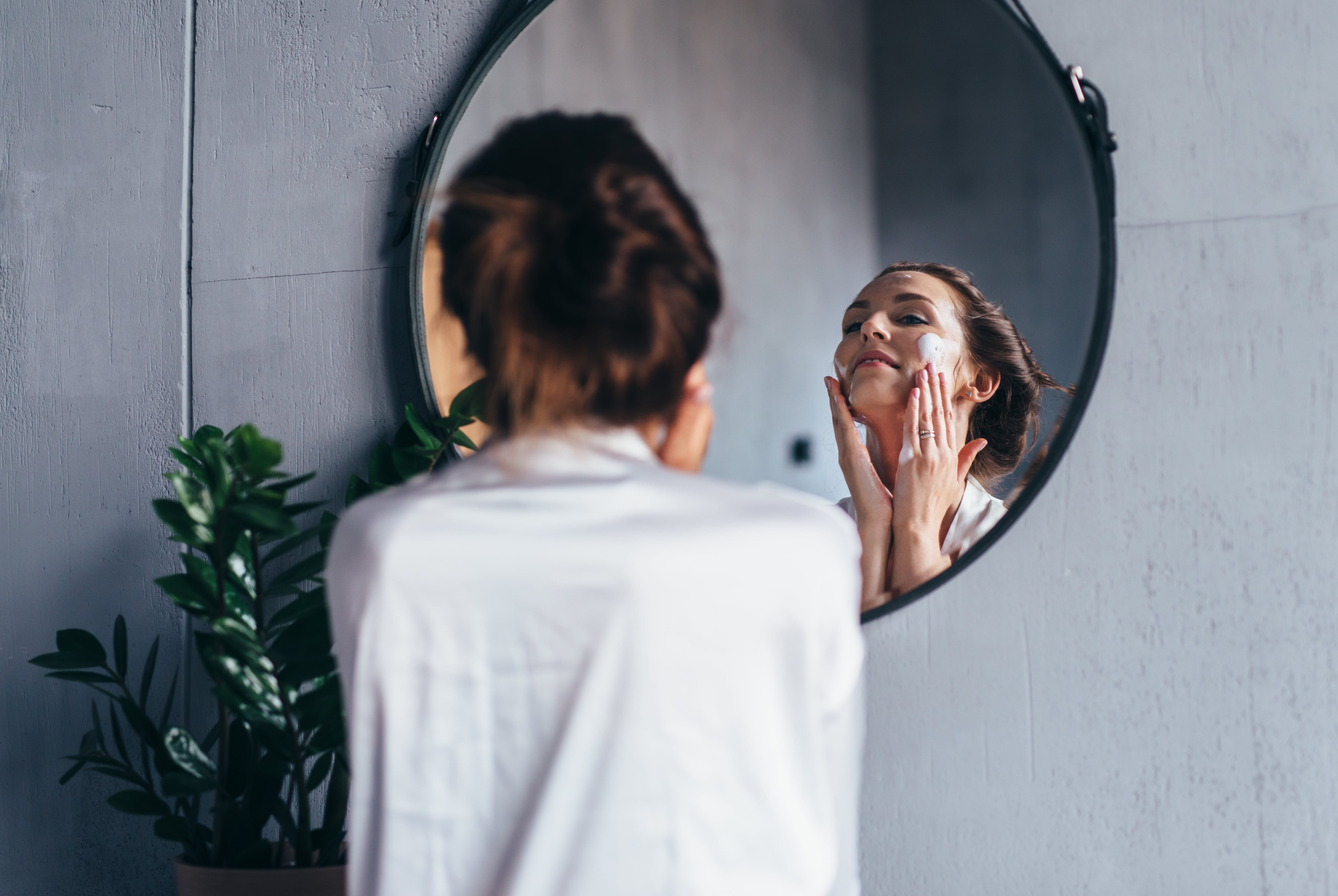 Frau blickt in einen runden Spiegel an einer grauen Wand. Sie trägt eine weiße Bluse, die braunen Haare zu einem Zopf gebunden und hat den Kopf in den Nacken gelegt. Mit den beiden Händen trägt sie Creme auf ihre Wangen auf.