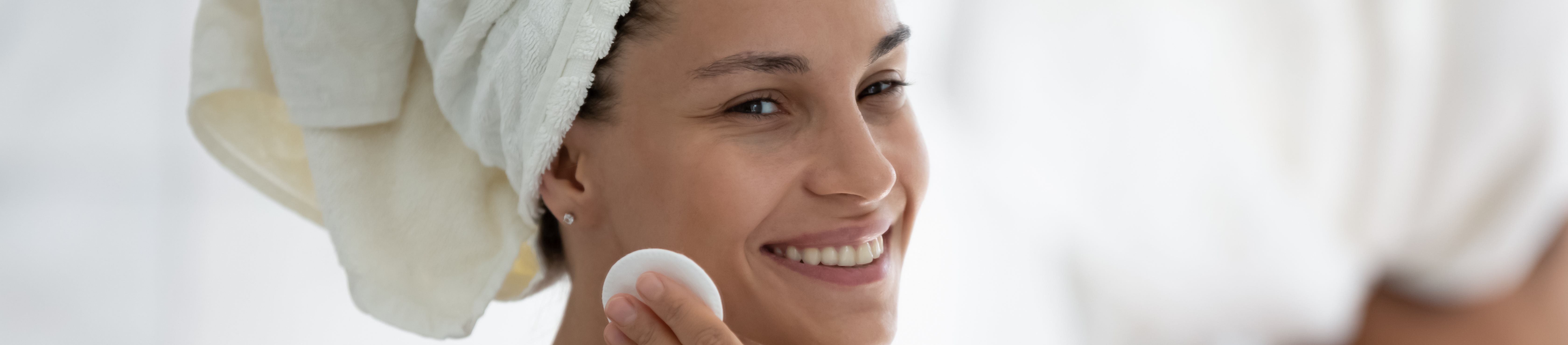 Eine lächelnde Frau, die ein Handtuch um den Kopf gewickelt hat, trägt mit einem Wattepad ein Hautpflegeprodukt auf ihren Hals auf.