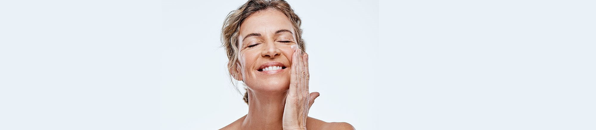 Donna matura sorride mentre applica una crema contorno occhi