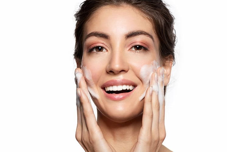 Ragazza sorride mentre massaggia il viso con un detergente schiumoso durante la doppia detersione quotidiana.