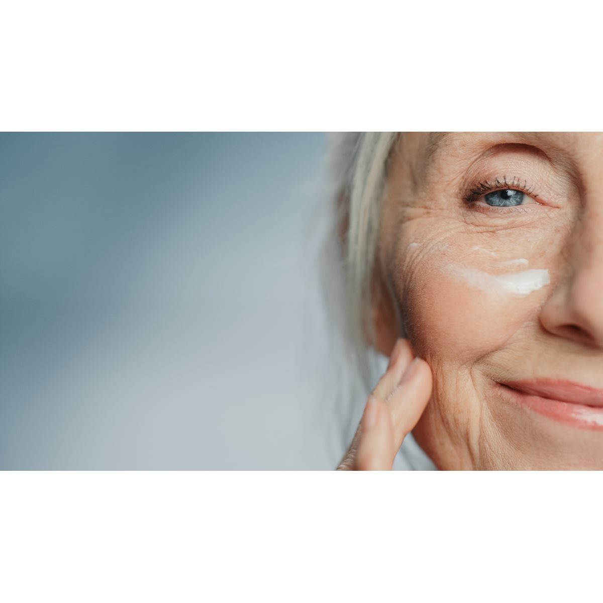 Une femme âgée à la peau éclatante et aux rides légères applique délicatement une crème hydratante sur son visage.