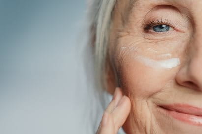 Eine ältere Frau mit strahlender Haut und leichten Falten trägt sanft eine Feuchtigkeitscreme auf ihr Gesicht auf.