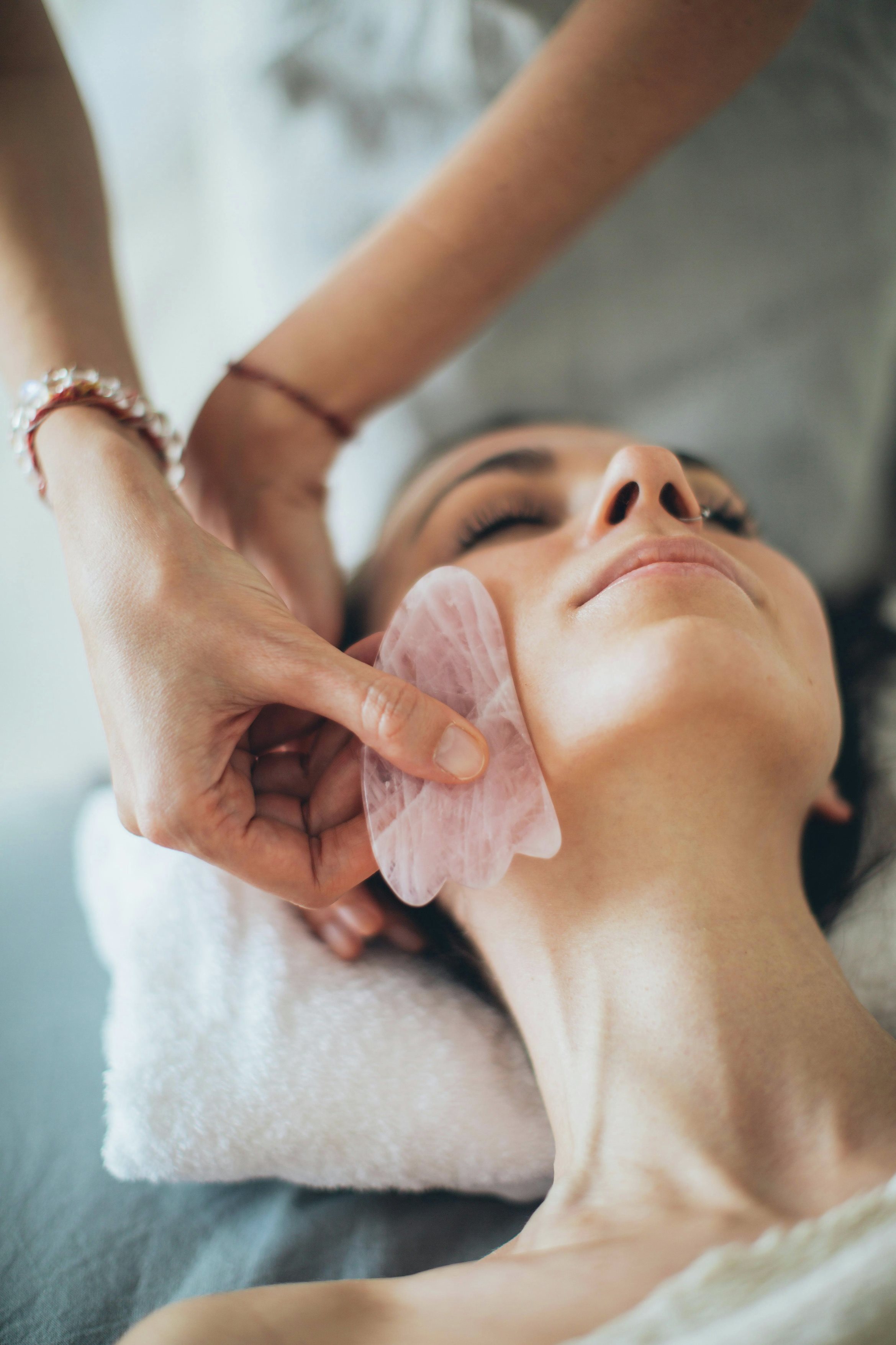 Nahaufnahme einer Gesichtsbehandlung mit rosafarbenem Gua Sha Stein entlang des Kiefers einer liegenden Frau.