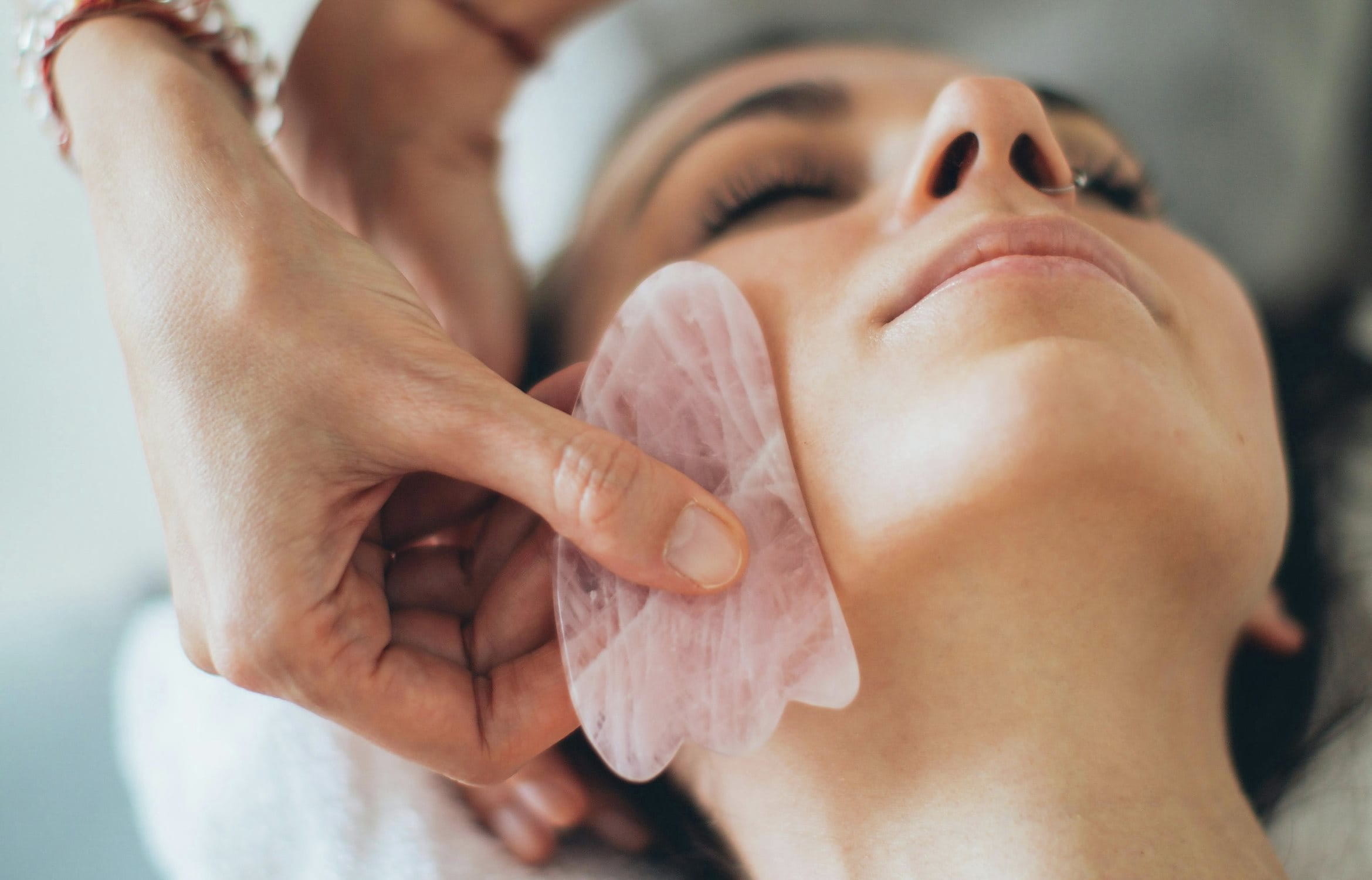 Nahaufnahme einer Gesichtsbehandlung mit rosafarbenem Gua Sha Stein entlang des Kiefers einer liegenden Frau.