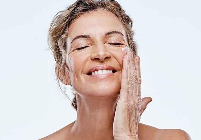 Donna matura sorride mentre applica una crema contorno occhi