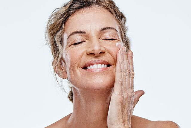 Donna matura sorride mentre applica una crema contorno occhi