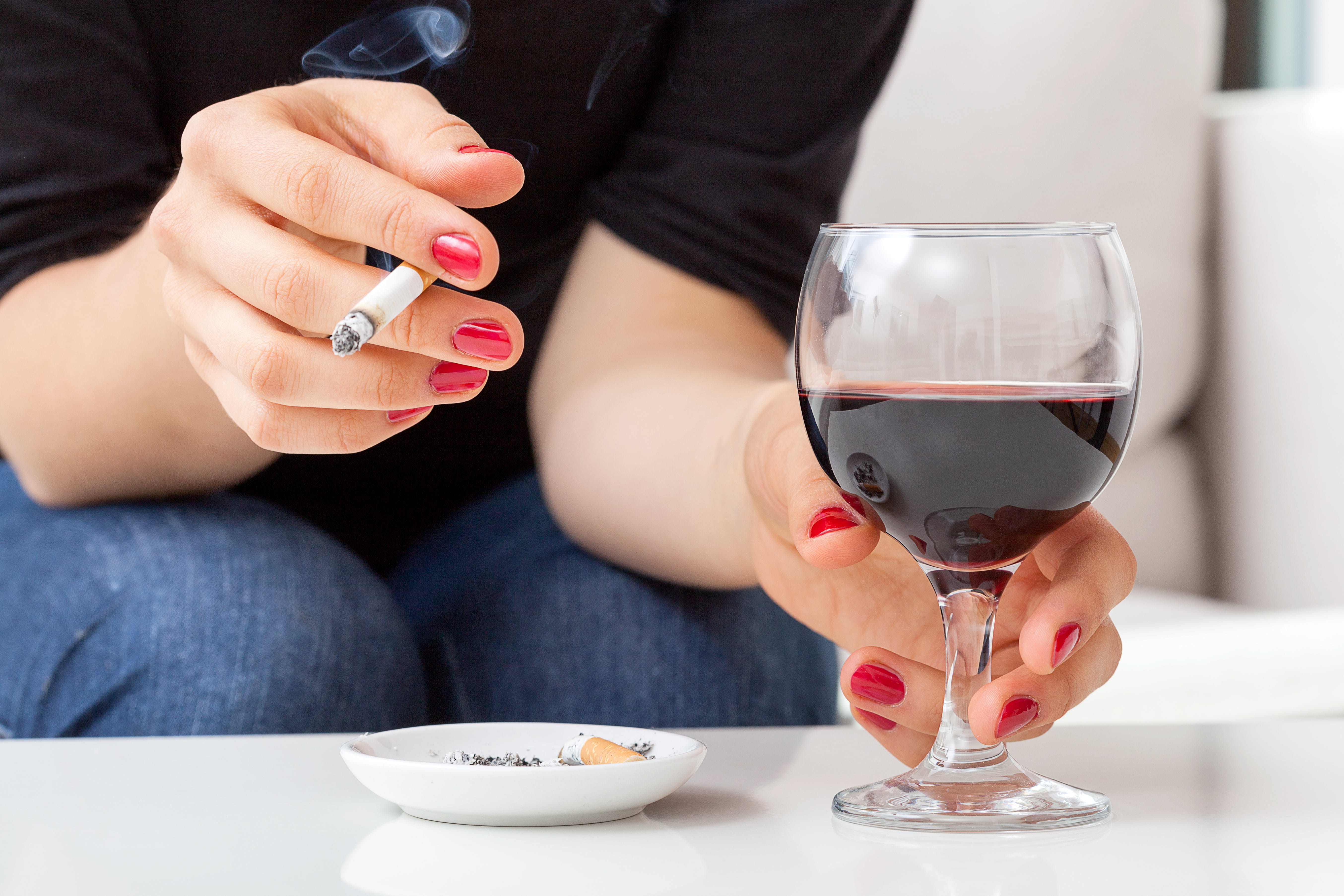 Eine Frau sitzt vor einem Tisch und hält in der einen Hand eine rauchende Zigarette und in der anderen ein Glas mit Rotwein.