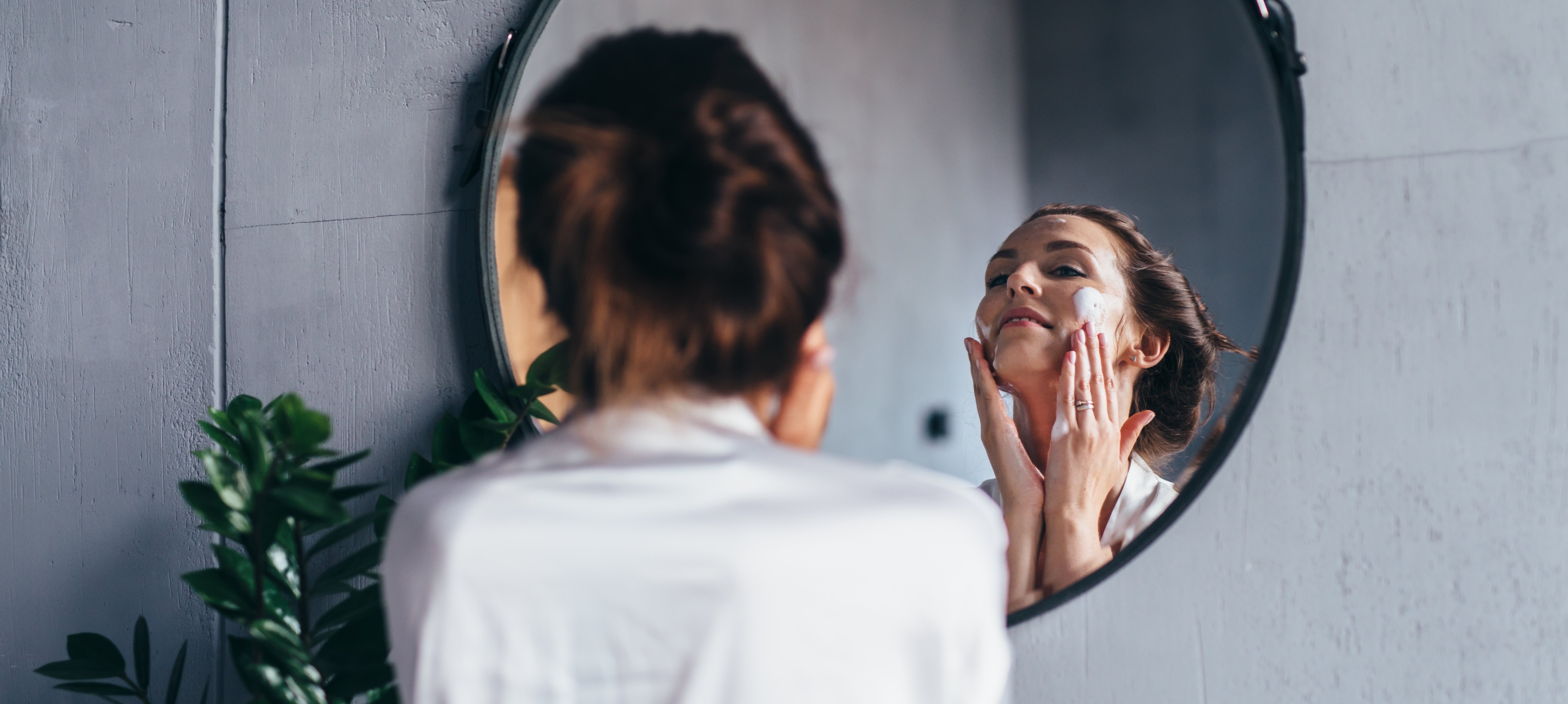 Frau blickt in einen runden Spiegel an einer grauen Wand. Sie trägt eine weiße Bluse, die braunen Haare zu einem Zopf gebunden und hat den Kopf in den Nacken gelegt. Mit den beiden Händen trägt sie Creme auf ihre Wangen auf.