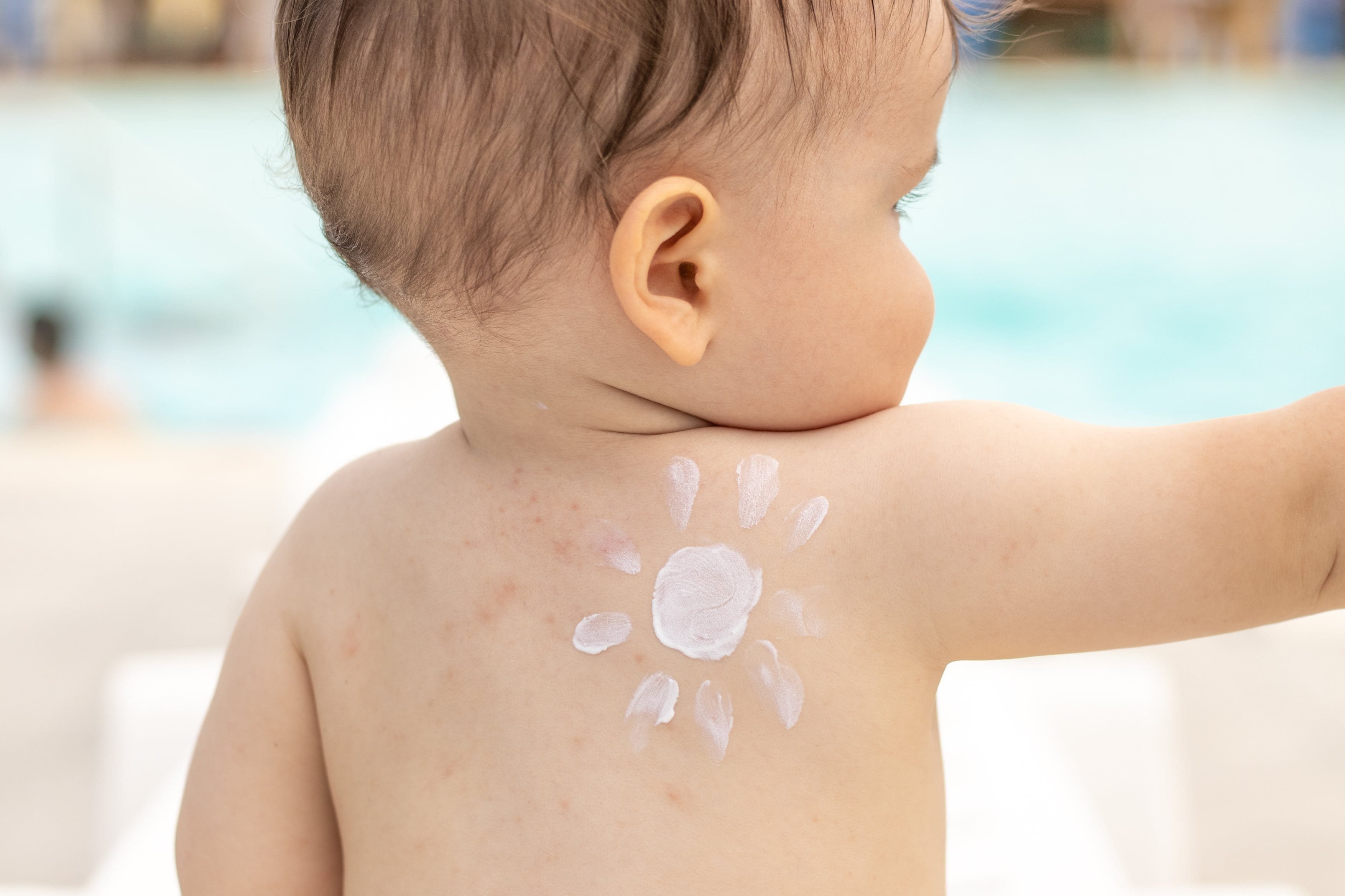  Ein Baby von hinten auf dessen Rücken eine Sonne aus Lotion gemalt wurde. Im Hintergrund ist Wasser zu sehen.
