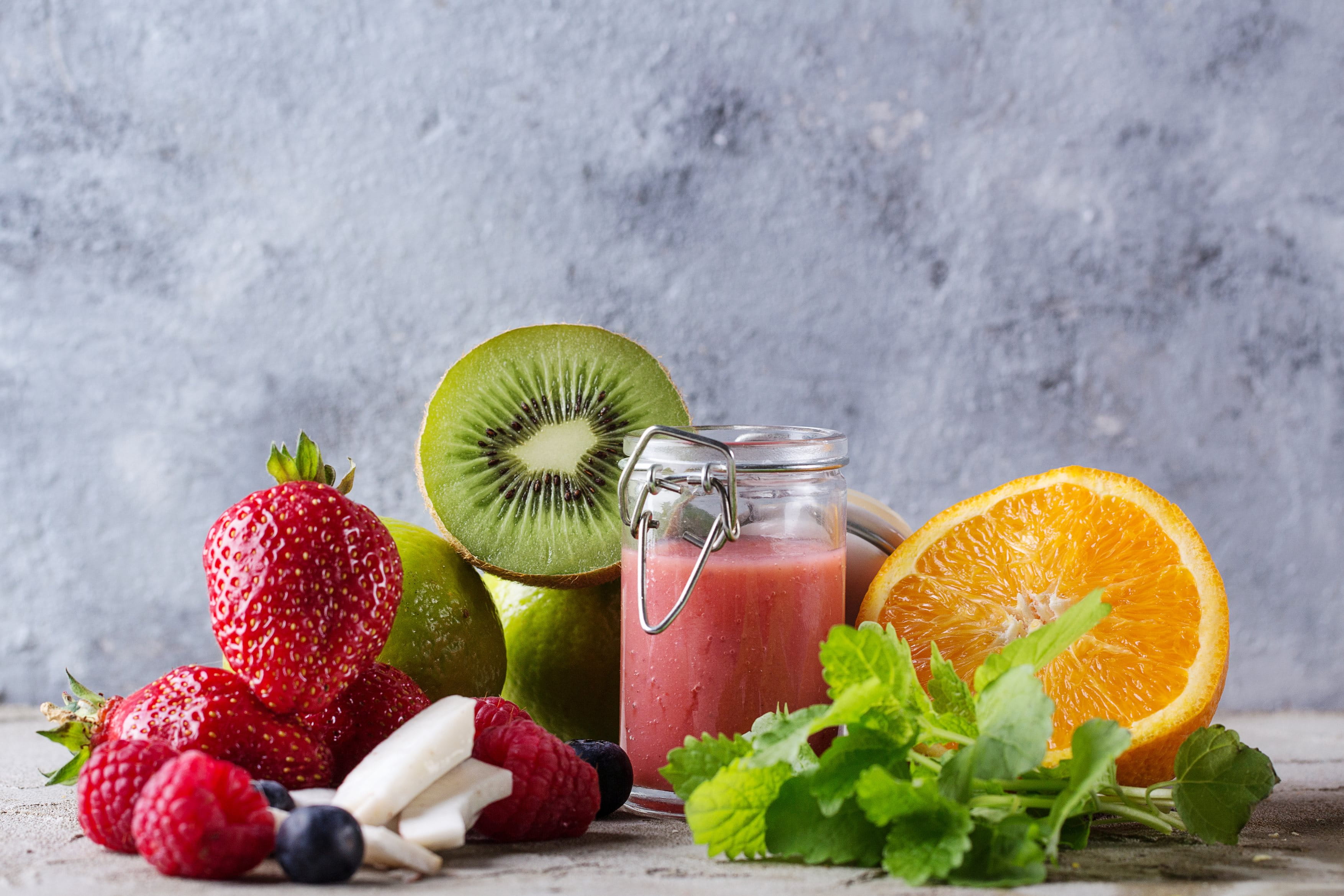 Eine graue Tischplatte, auf der Erbeeren, Kiwis, Blaubeeren, Kokosnussstücke, ein Beeren-Smoothie sowie eine aufgeschnittene orange und Minze zu sehen sind.