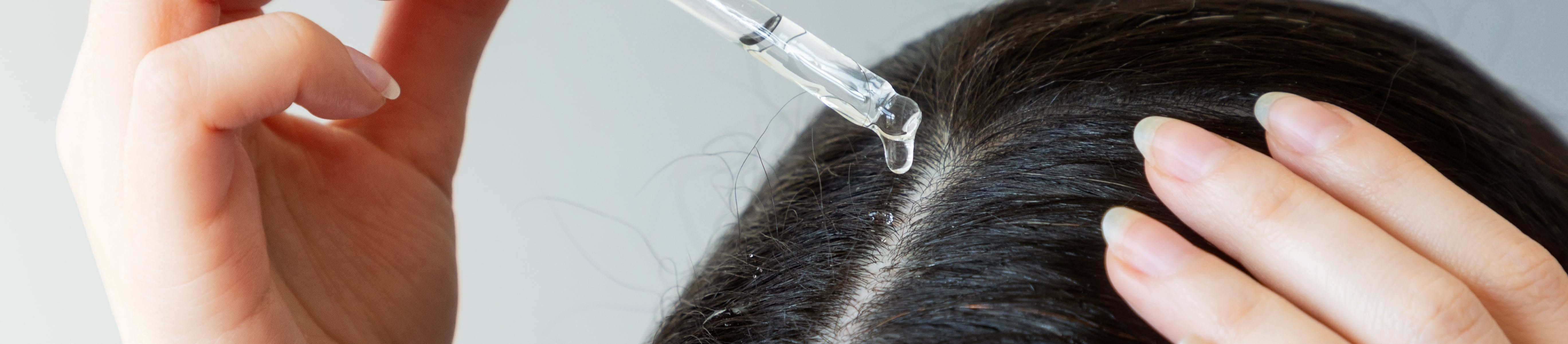 Nahaufnahme des Kopfes einer Frau, die mit der rechten Hand aus einer Pipette Öl auf ihre dunklen Haare aufträgt.