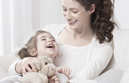 Little girl, laughing and holding a stuffed animal is sitting on her mom´s lap.