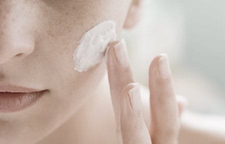 Woman is applying cream on her left cheek with finger.