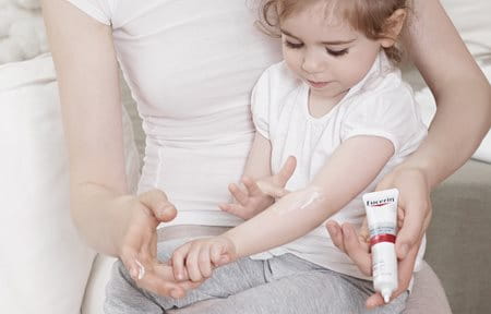 Little girl, sitting on mother´s lap, is applying Eucerin AtopiControl Acute Care cream on the crook of her arm.