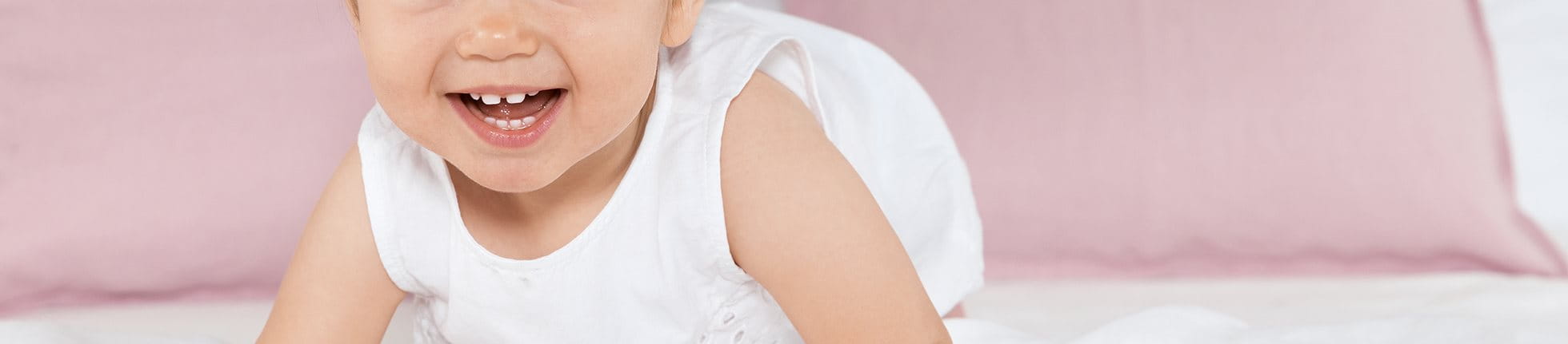 Smiling baby with atopic dermatitis in white dress on bed.