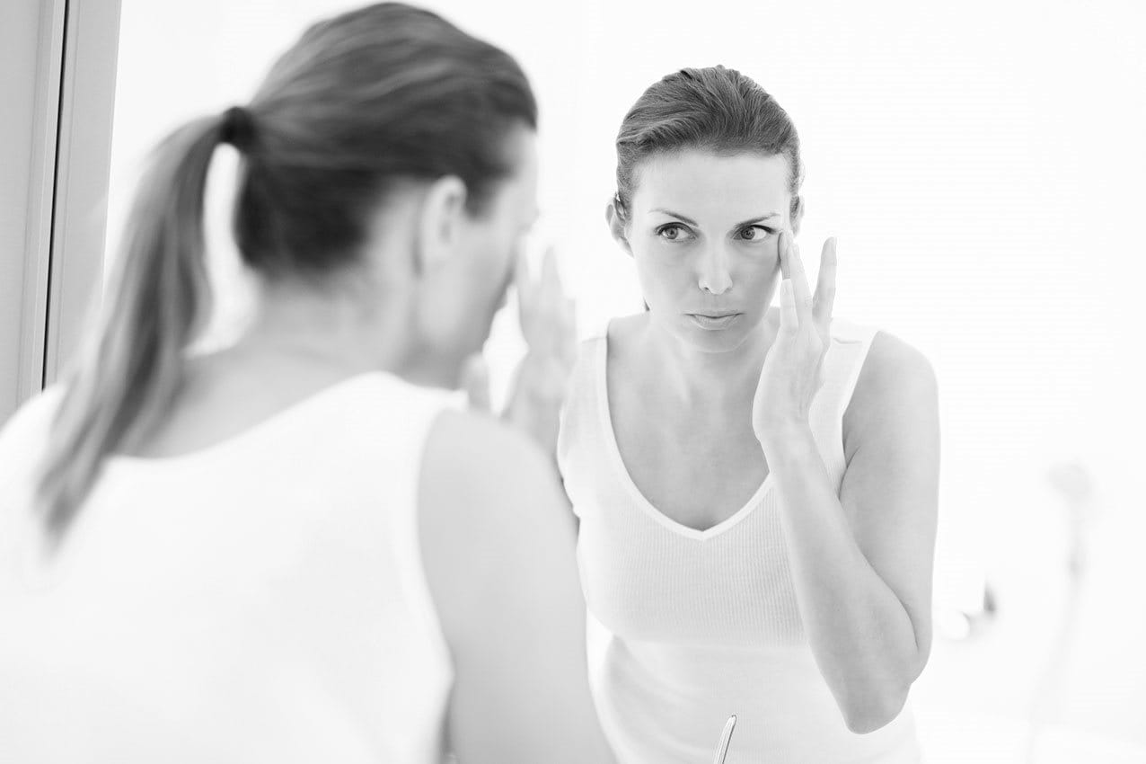 Frau betrachtet sich im Spiegel, greift mit Finger an die Schläfe