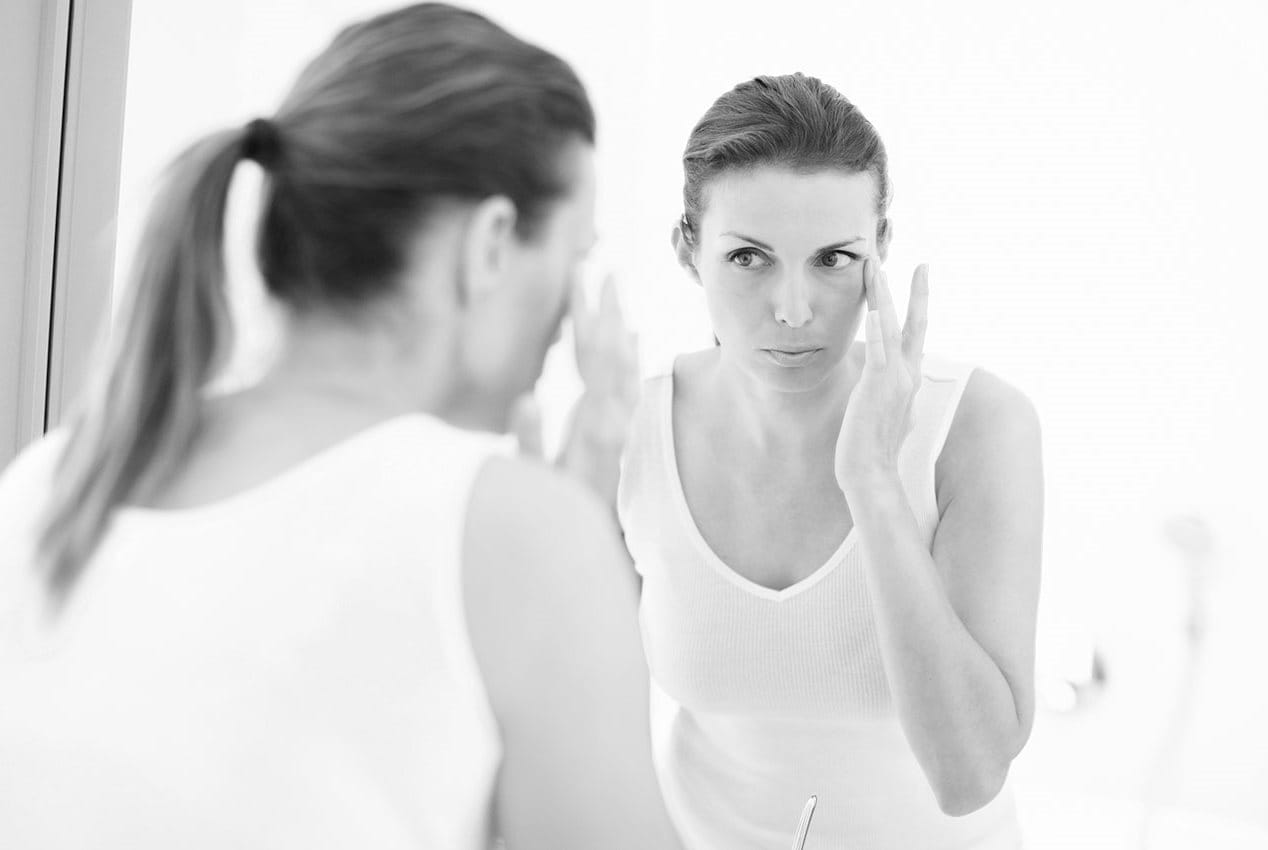 Frau betrachtet sich im Spiegel, greift mit Finger an die Schläfe