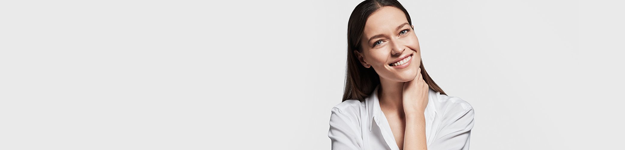 A front view of a female model with brown hair and light coloured eyes smiling while holding the left side of her neck.