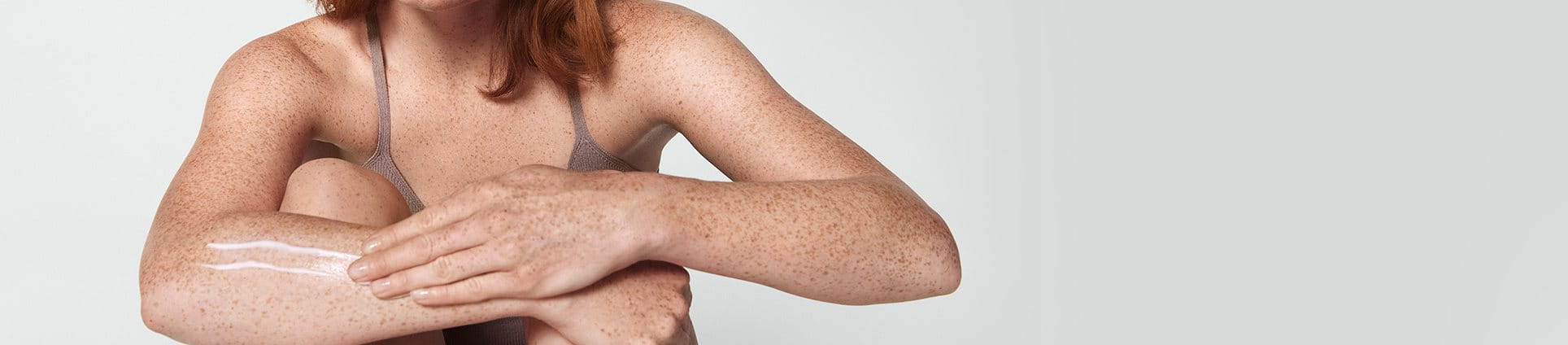 View of a female model with short red hair smearing a cream product along her right arm.