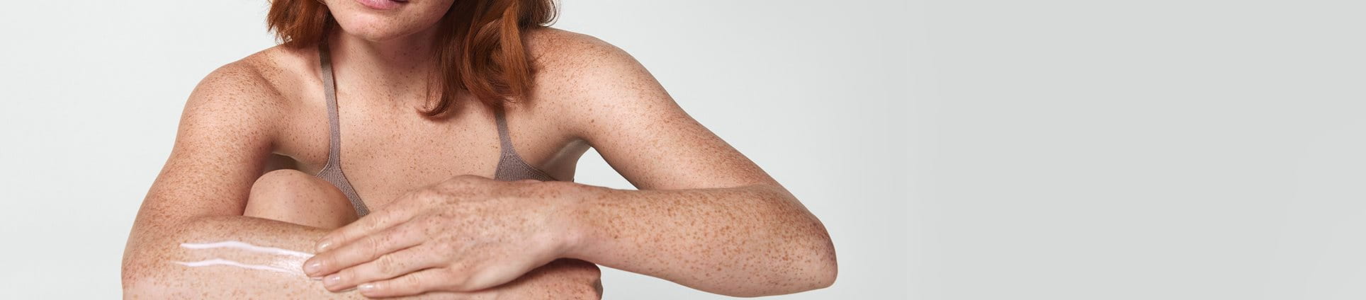 View of a female model with short red hair smearing a cream product along her right arm.