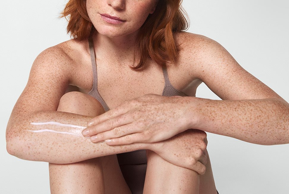 Une femme mannequin aux cheveux roux courts  étend une touche de produit en crème sur son bras droit.