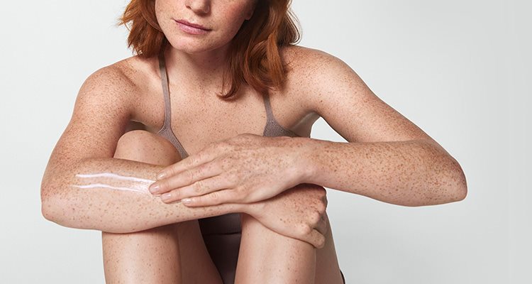 View of a female model with short red hair smearing a cream product along her right arm.