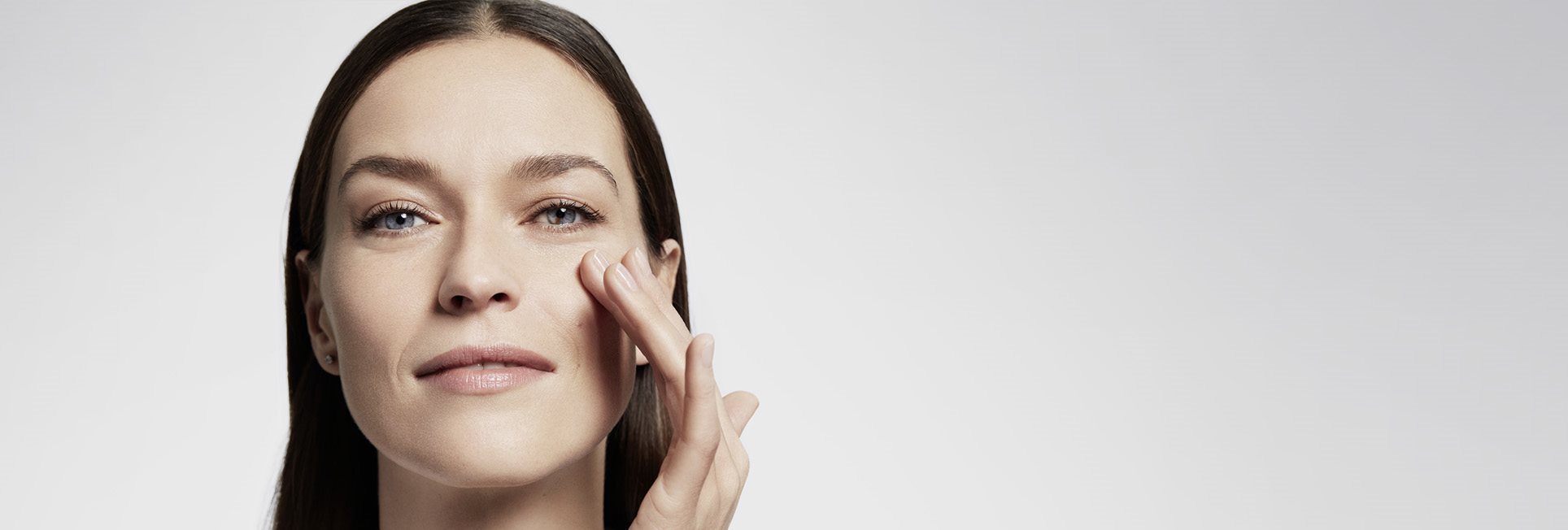 A closeup view of a female model with brown hair and blue eyes touching her left cheek and slightly smiling.
