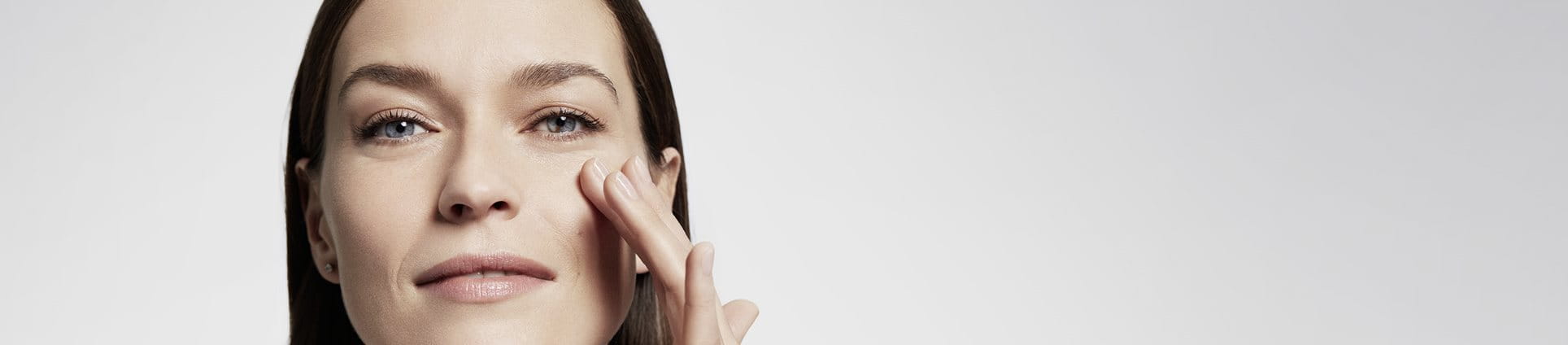 Plan rapproché d'une femme mannequin aux cheveux bruns et aux yeux bleus, touchant sa joue gauche tout en esquissant un sourire.