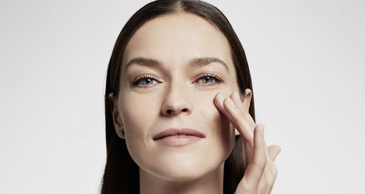 Plan rapproché d'une femme mannequin aux cheveux bruns et aux yeux bleus, touchant sa joue gauche tout en esquissant un sourire.