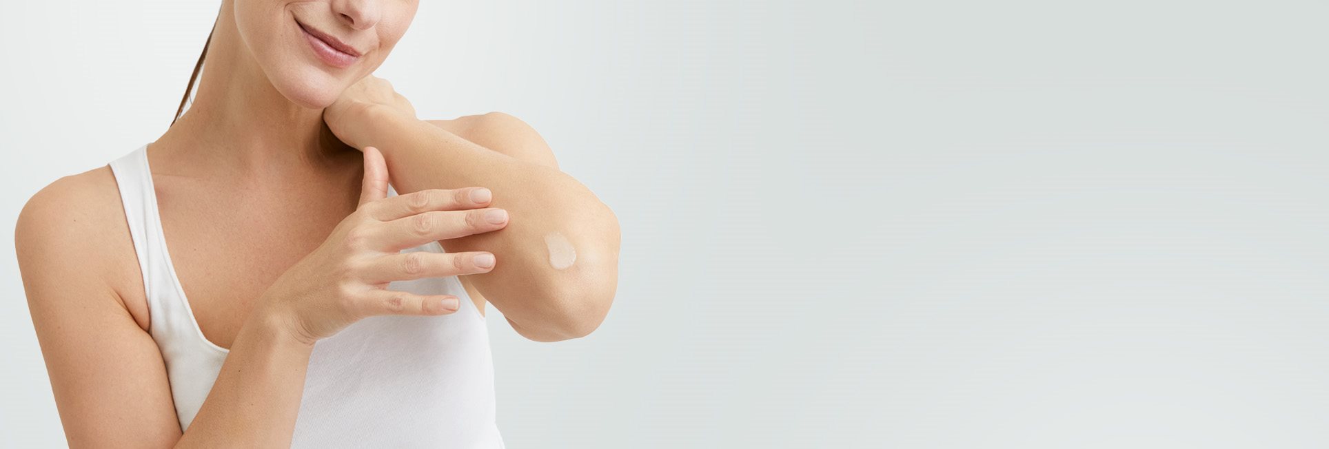 View of a female model smiling with a cream product smudged on her left elbow against a grey background.