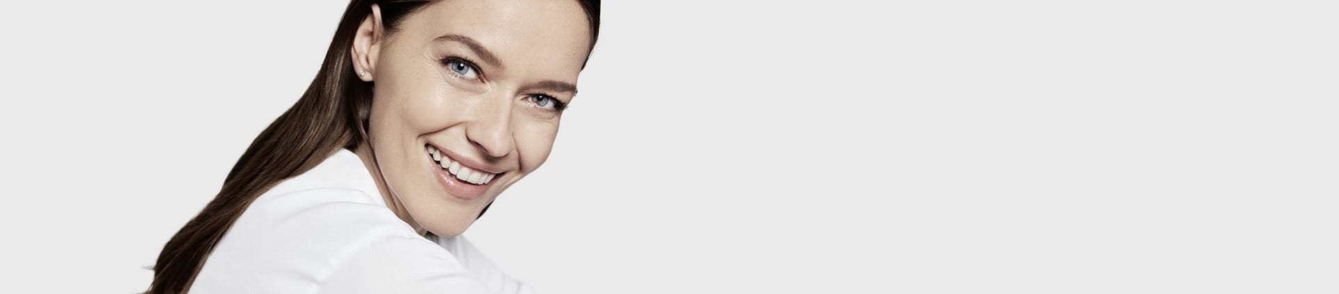 View of a female model with brown hair and blue eyes smiling while wearing a white coloured top against a white background.