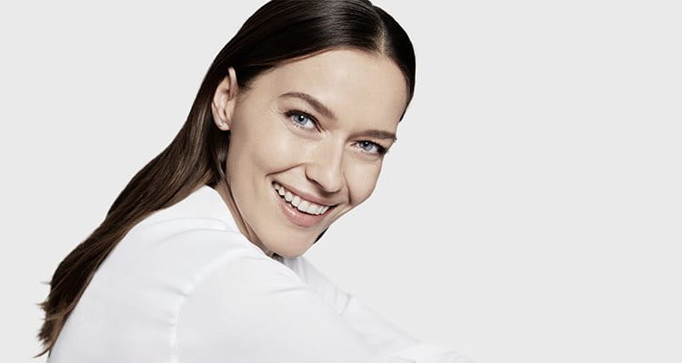 View of a female model with brown hair and blue eyes smiling while wearing a white coloured top against a white background.