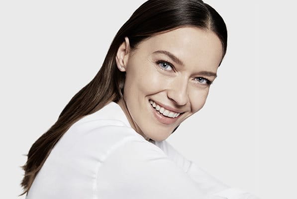 View of a female model with brown hair and blue eyes smiling while wearing a white coloured top against a white background.