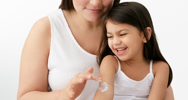 A closeup view of a female model and a little girl smiling with a cream product being applied to her right arm.