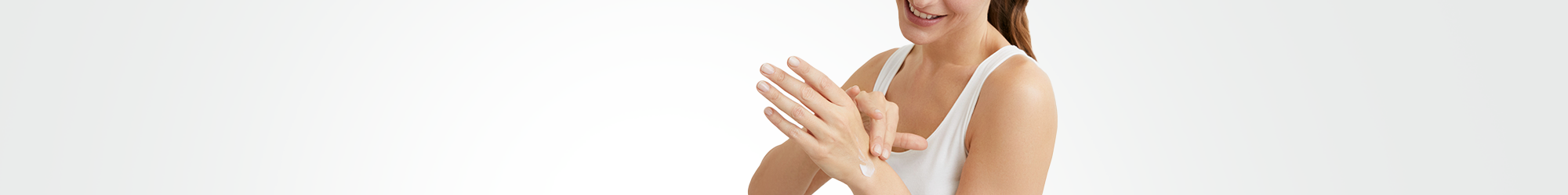 View of a female model wearing a white tank top and applying a cream product to her left hand.