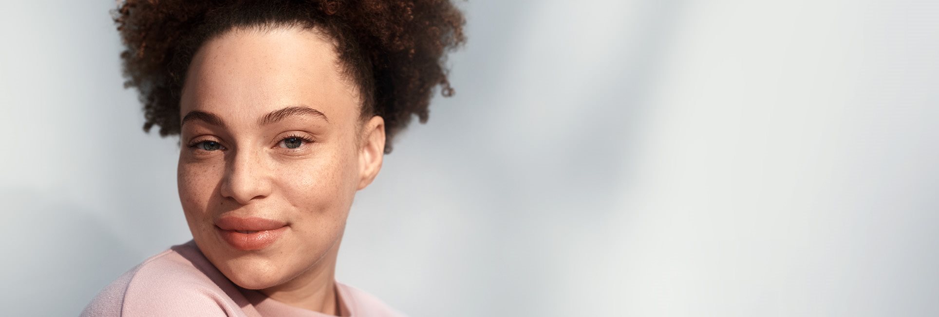  Vue d&rsquo;un mannequin féminin aux cheveux bouclés foncés relevés en queue de cheval et portant un haut de couleur blush, souriant sur un fond gris.