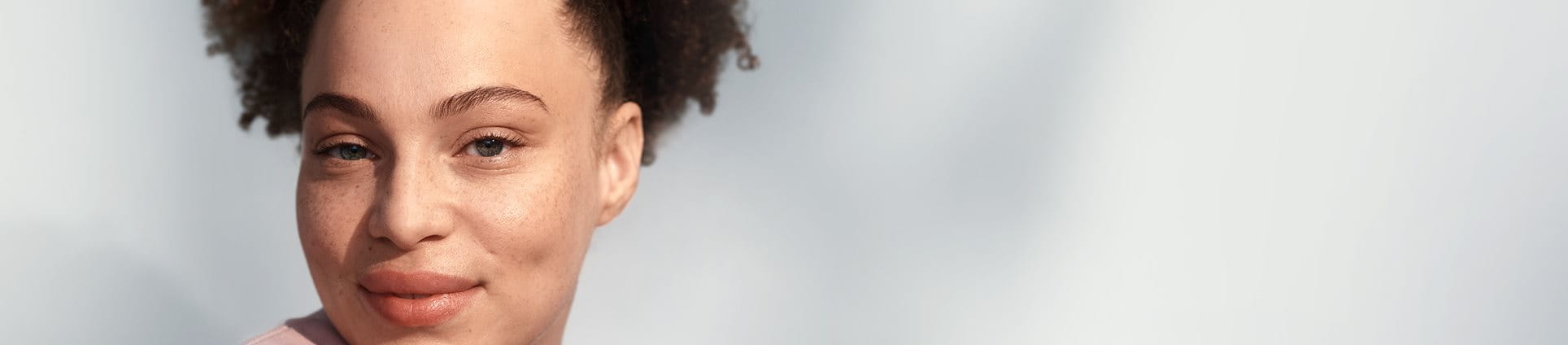 Vue d’un mannequin féminin aux cheveux bouclés foncés relevés en queue de cheval et portant un haut de couleur blush, souriant sur un fond gris.