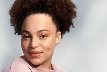 A view of a female model with dark curly hair pulled up into a ponytail and wearing a blush coloured top while smiling against a grey background.